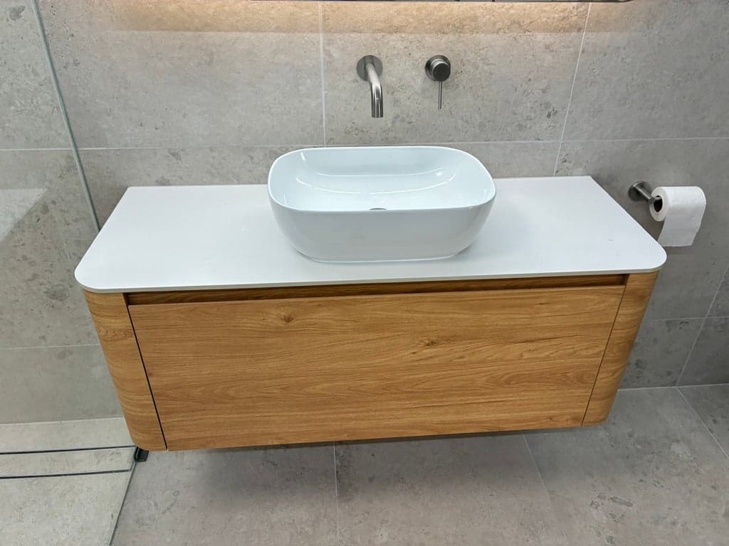 A floating timber vanity with a white top, vessel basin and wall-mounted tap against large grey tiles